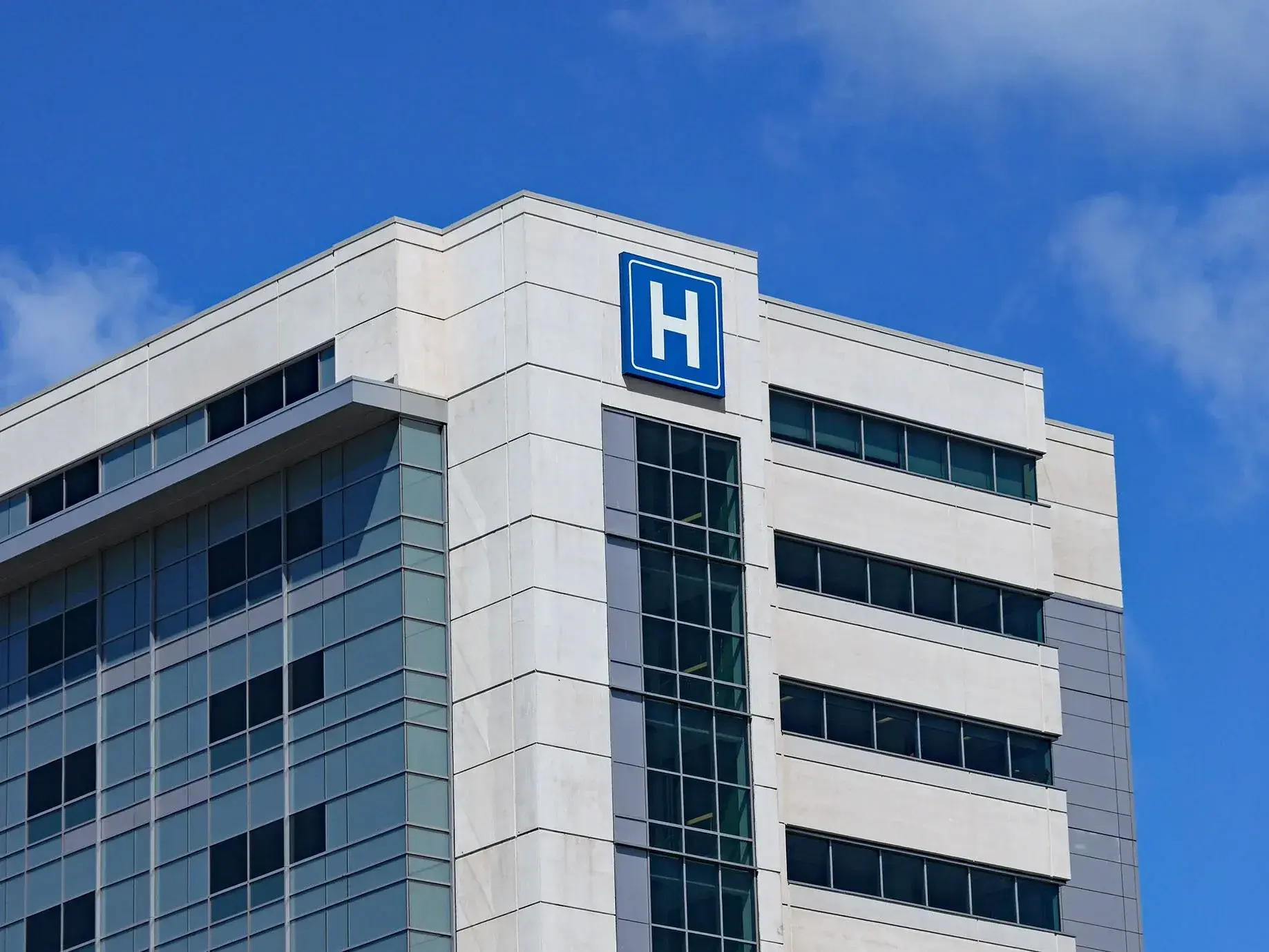 Een modern ziekenhuisgebouw met een wit en glasgevelontwerp, gefotografeerd tegen een blauwe lucht met enkele wolken. Op de bovenhoek van het gebouw is een blauw bord met een witte letter "H" zichtbaar, wat duidt op een ziekenhuis of medische faciliteit.