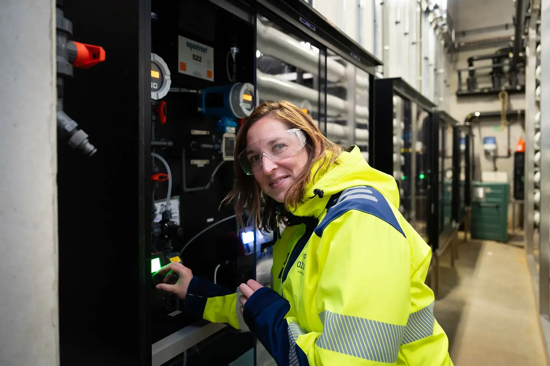 Een vrouw met halflang bruin haar en een veiligheidsbril bedient een controlepaneel in een industriële omgeving. Ze draagt een gele veiligheidsjas met reflecterende strepen en het logo van "azulatis". Op de achtergrond zijn rijen buizen, meters en technische installaties zichtbaar. Ze kijkt glimlachend naar de camera terwijl ze een knop indrukt.