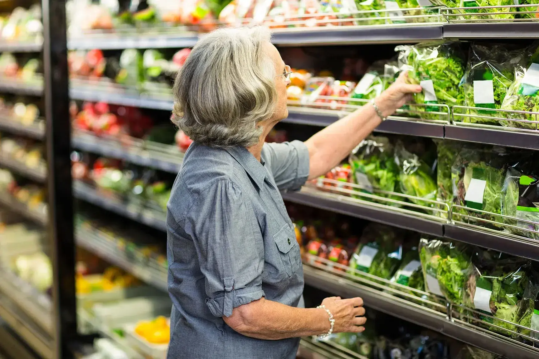 Een oudere vrouw met grijs haar en een bril winkelt in de groenteafdeling van een supermarkt. Ze draagt een blauwgrijze blouse en reikt naar een zak verse sla op een schap, terwijl er op de achtergrond meer verse producten zoals groenten en fruit zichtbaar zijn.