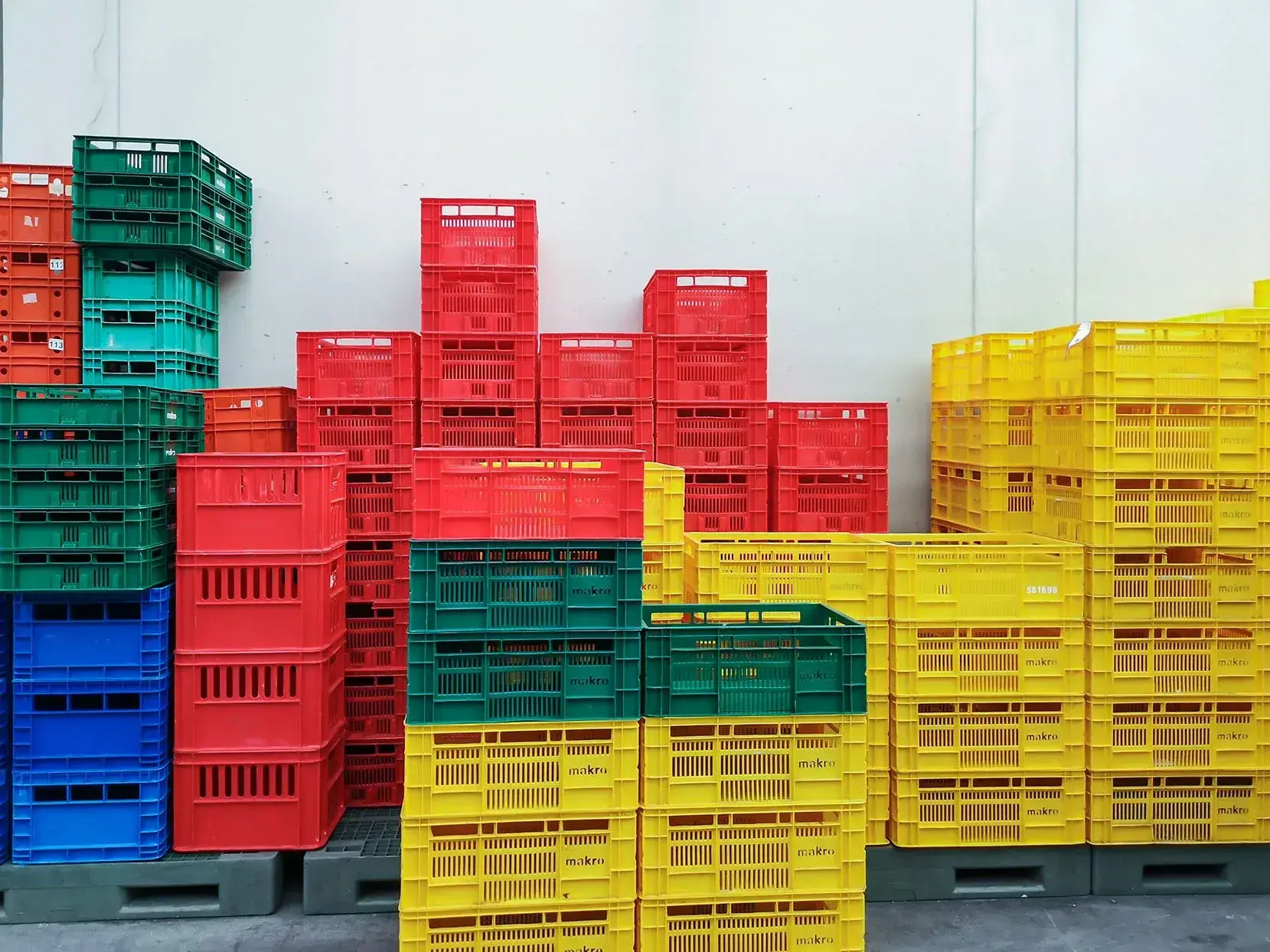Een stapel kleurrijke kunststof kratten in rood, geel, groen en blauw, netjes opgestapeld tegen een witte muur. De kratten, die vaak worden gebruikt voor transport en opslag van verse producten, staan op pallets in een georganiseerde indeling.