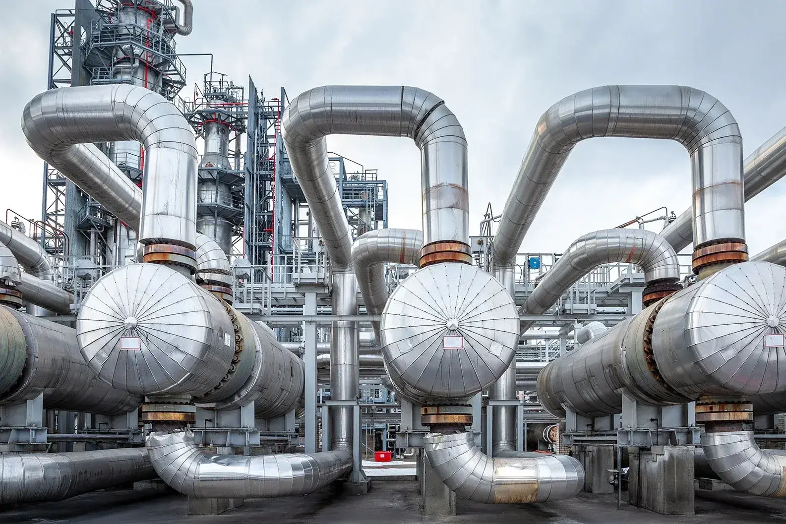 Een industriële installatie met een complex netwerk van grote, zilverkleurige pijpleidingen die in verschillende richtingen buigen. De metalen buizen zijn voorzien van ronde uiteinden en verbonden met kleppen en ondersteunende structuren. Op de achtergrond zijn extra leidingen en staalconstructies zichtbaar tegen een bewolkte lucht.
