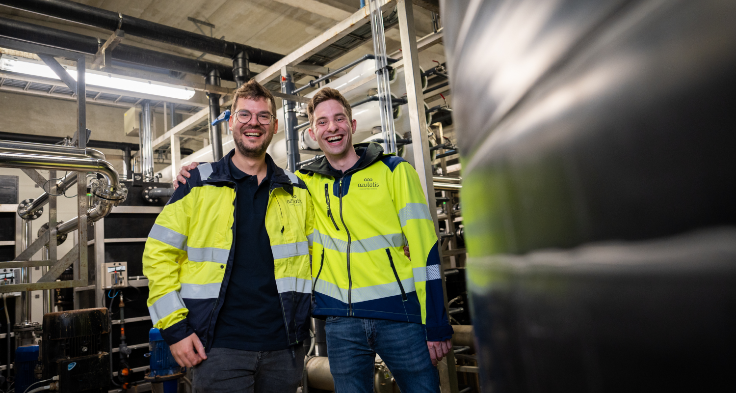 Twee jonge mannen met brillen en lichte baarden staan lachend naast elkaar in een industriële omgeving, omringd door metalen buizen en machines. Ze dragen gele veiligheidsjassen met reflecterende strepen en hebben een ontspannen houding, met een arm om elkaars schouder. De achtergrond toont een technische installatie met leidingen en structuren.