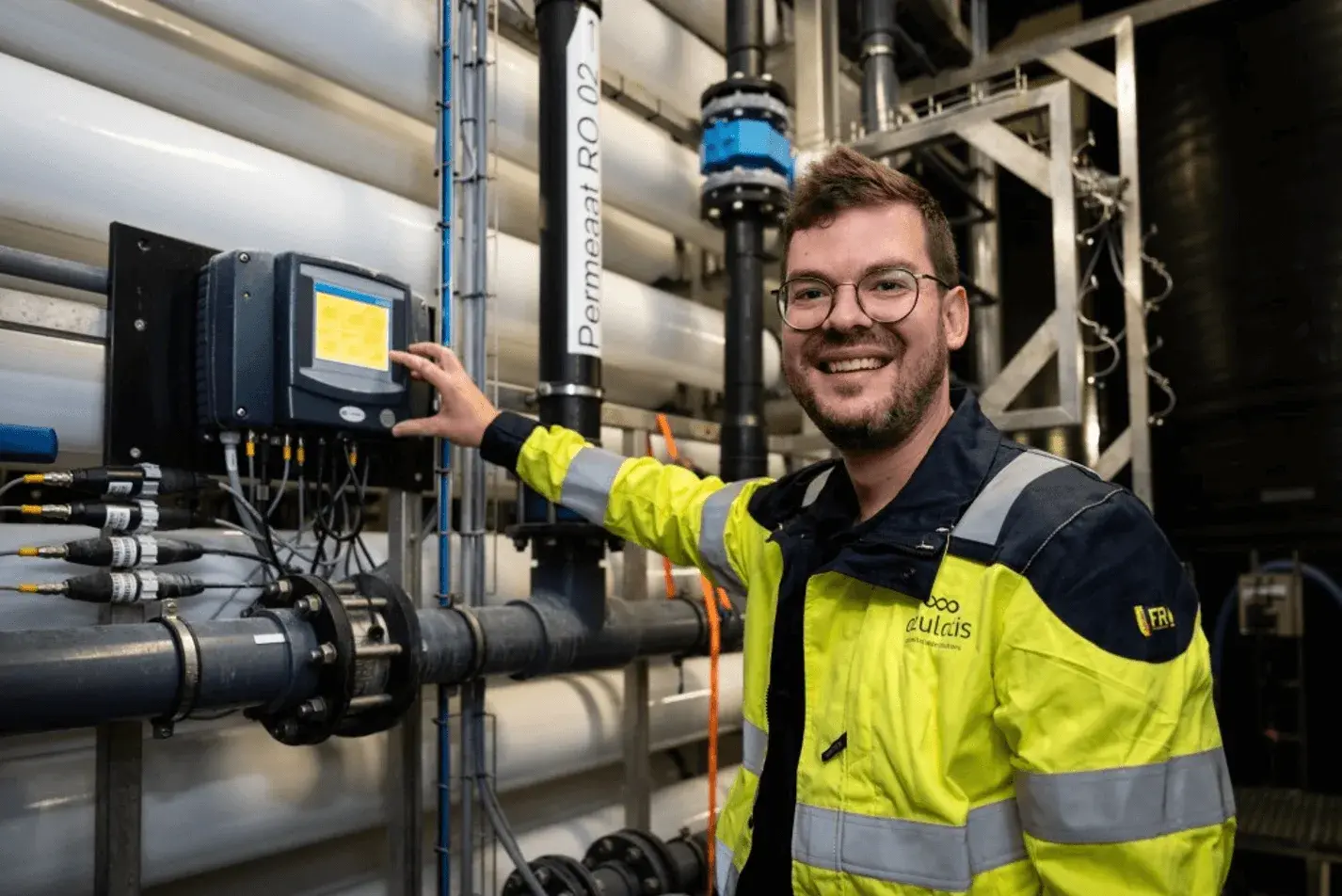 Een lachende man met een bril en een gele veiligheidsjas bedient een bedieningspaneel in een industriële waterzuiveringsinstallatie. Op de achtergrond zijn grote witte buizen, leidingen en technische apparatuur zichtbaar. De omgeving heeft een moderne en functionele uitstraling.