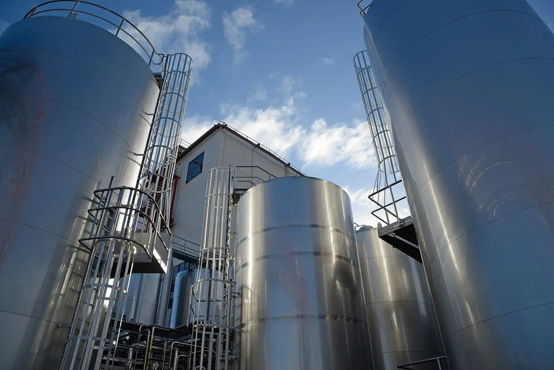 Industriële opslagtanks van roestvrij staal, gefotografeerd vanuit een laag perspectief. De tanks zijn voorzien van metalen trappen en veiligheidsleuningen. Op de achtergrond is een fabriekspand zichtbaar, met een blauwe lucht en enkele wolken erboven.